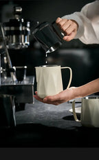 Black stainless milk pitcher pouring into a white cup, angled for latte art