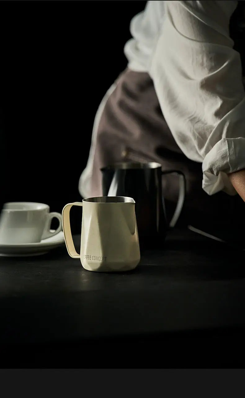 Milky white stainless milk pitcher with handle, beside a black pitcher for latte art
