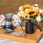 Outdoor pour-over coffee setup with glass dripper, gooseneck kettle, mug, and filter pack