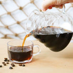 Stainless kettle pouring hot coffee into glass cup, with beans nearby from pour-over kit