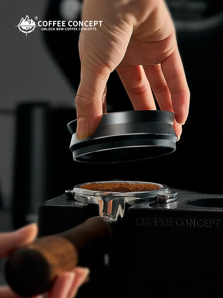 Black magnetic espresso dosing ring lifted by hand over a portafilter basket with grounds.