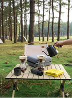 Pour-over coffee kit on outdoor table with kettle, dripper, and stainless mugs