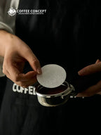 Hand holding a white circular coffee filter paper disc over a portafilter basket