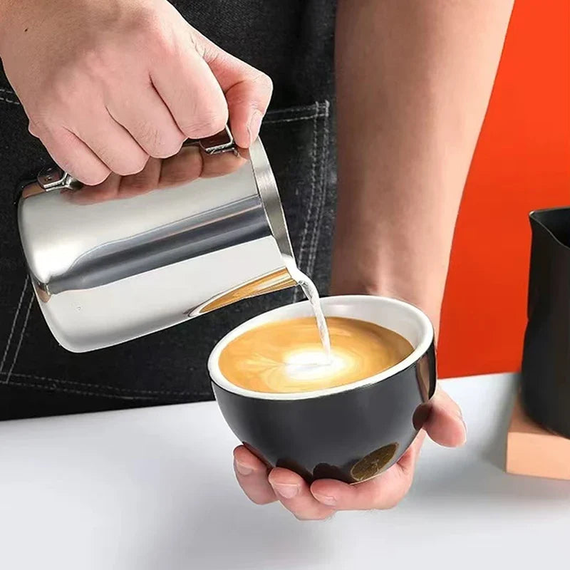 Stainless steel milk frothing pitcher pouring steamed milk into a coffee cup