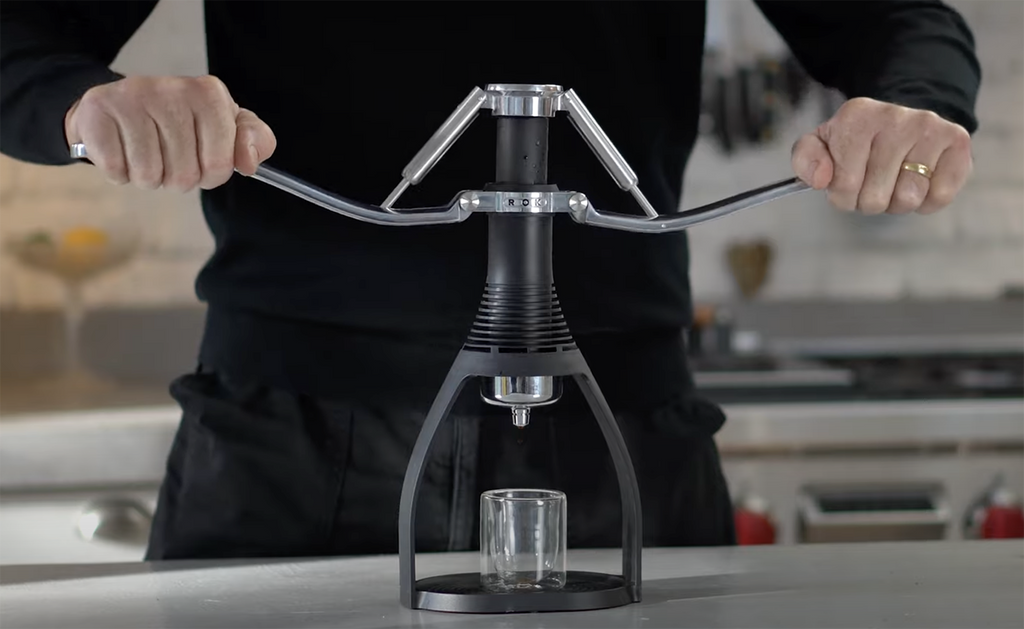 Black manual espresso maker being operated with hands, stainless handle assembly